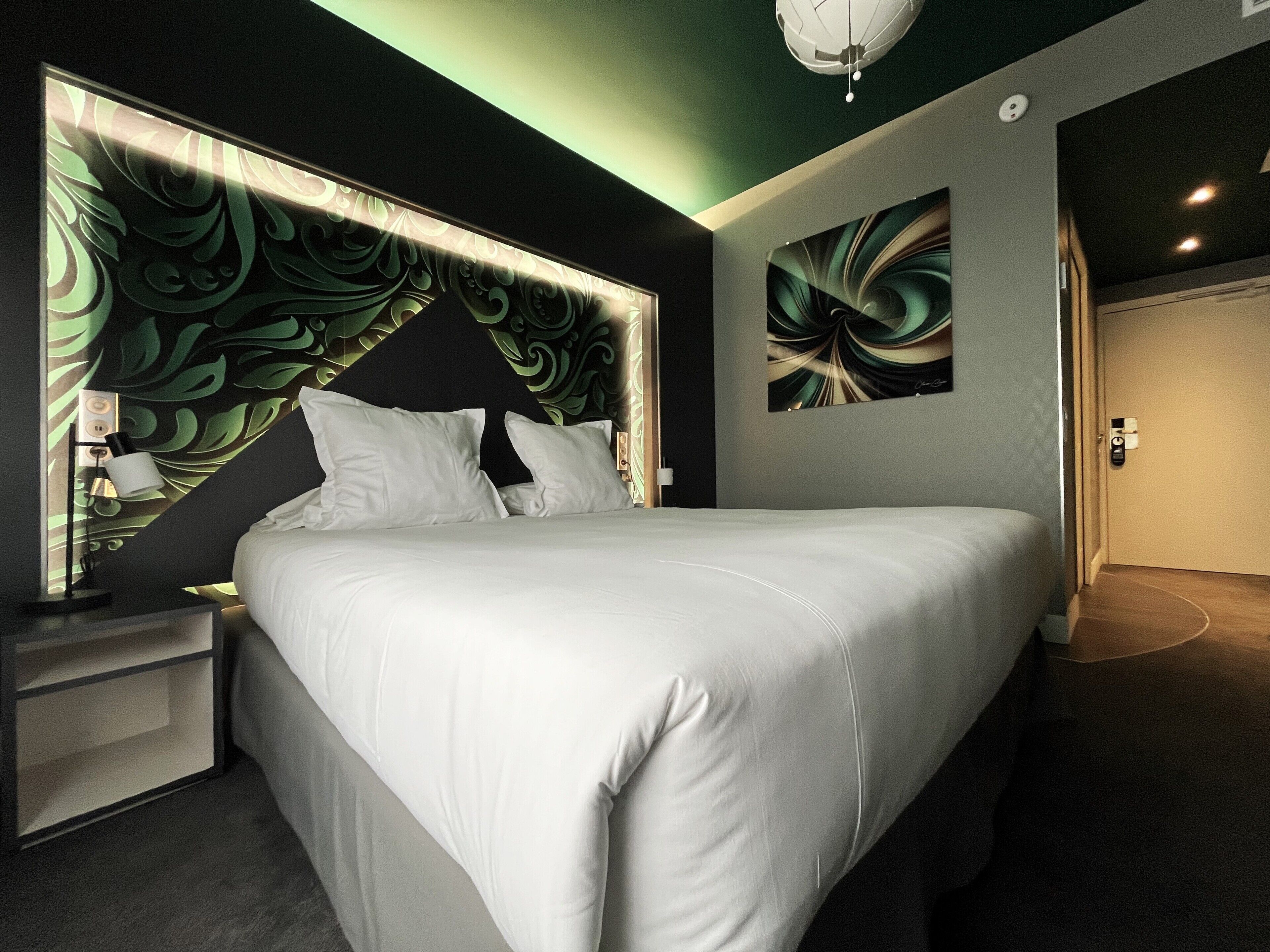 A modern bedroom with a large bed, a nightstand, a wall-mounted light, and a door.