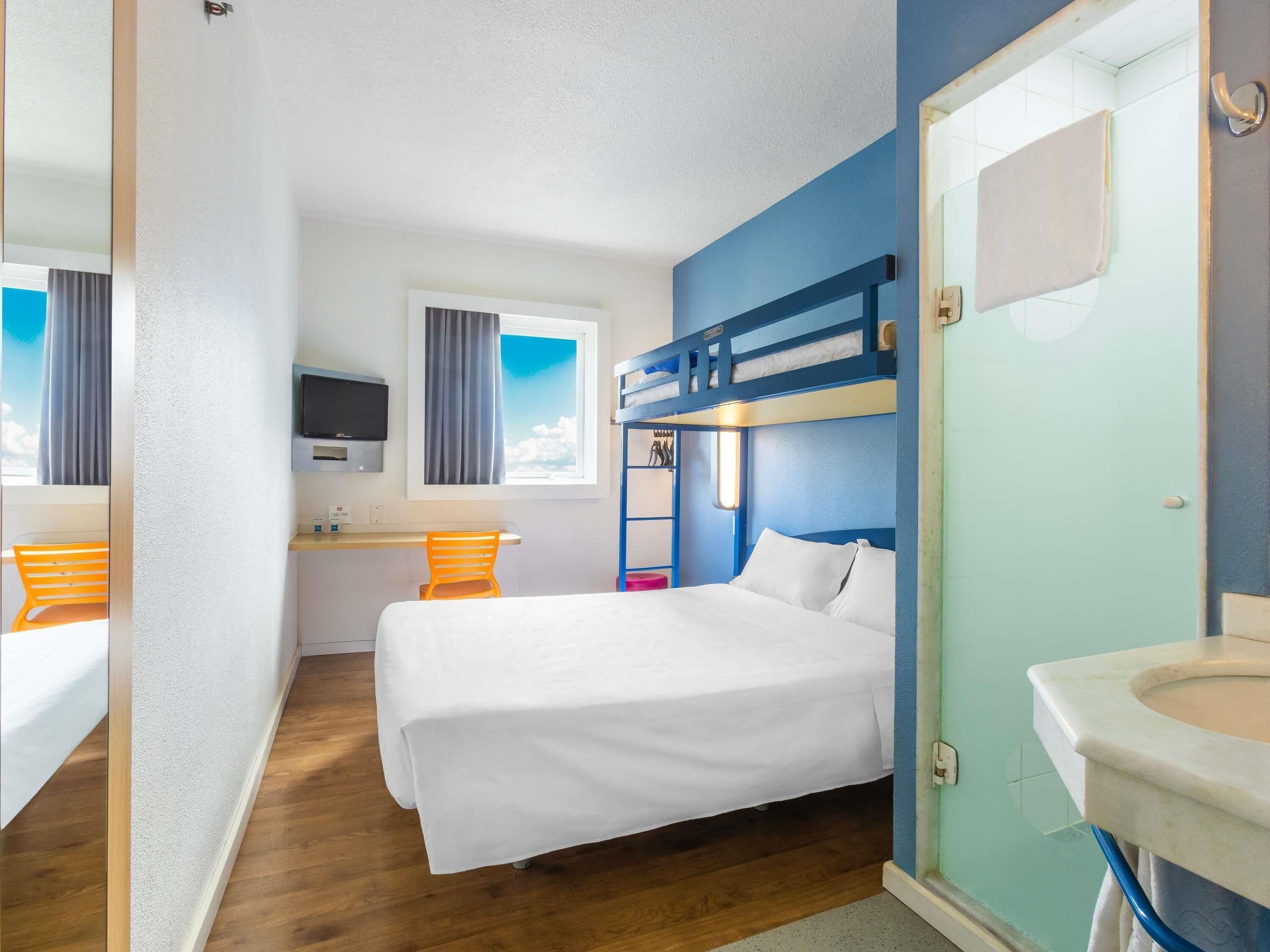 Una habitación con una cama litera, un escritorio con silla, televisión y un baño con lavamanos y espejo.