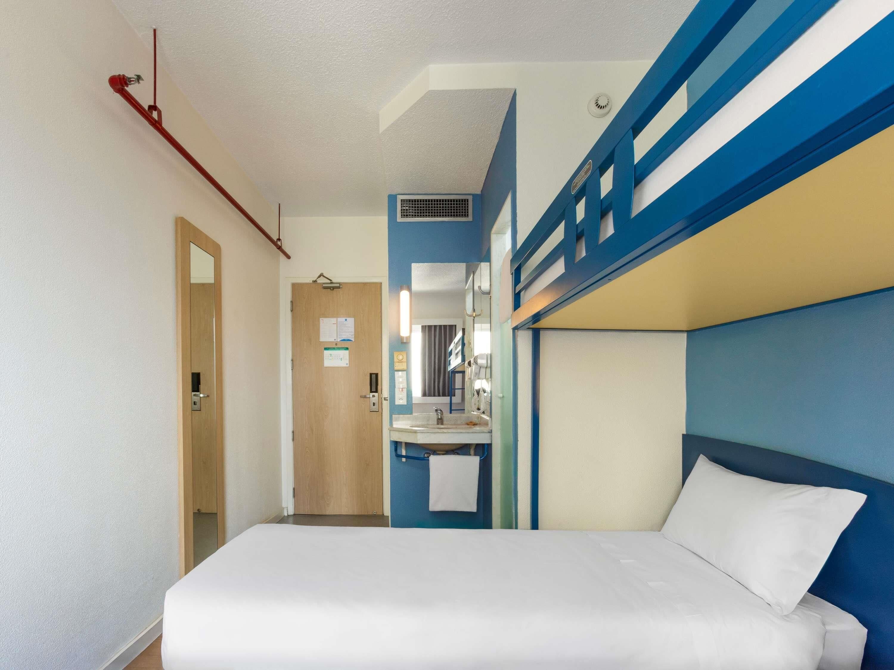 Habitación de hotel con cama, baño con lavabo y espejo, puerta y sistema de ventilación.