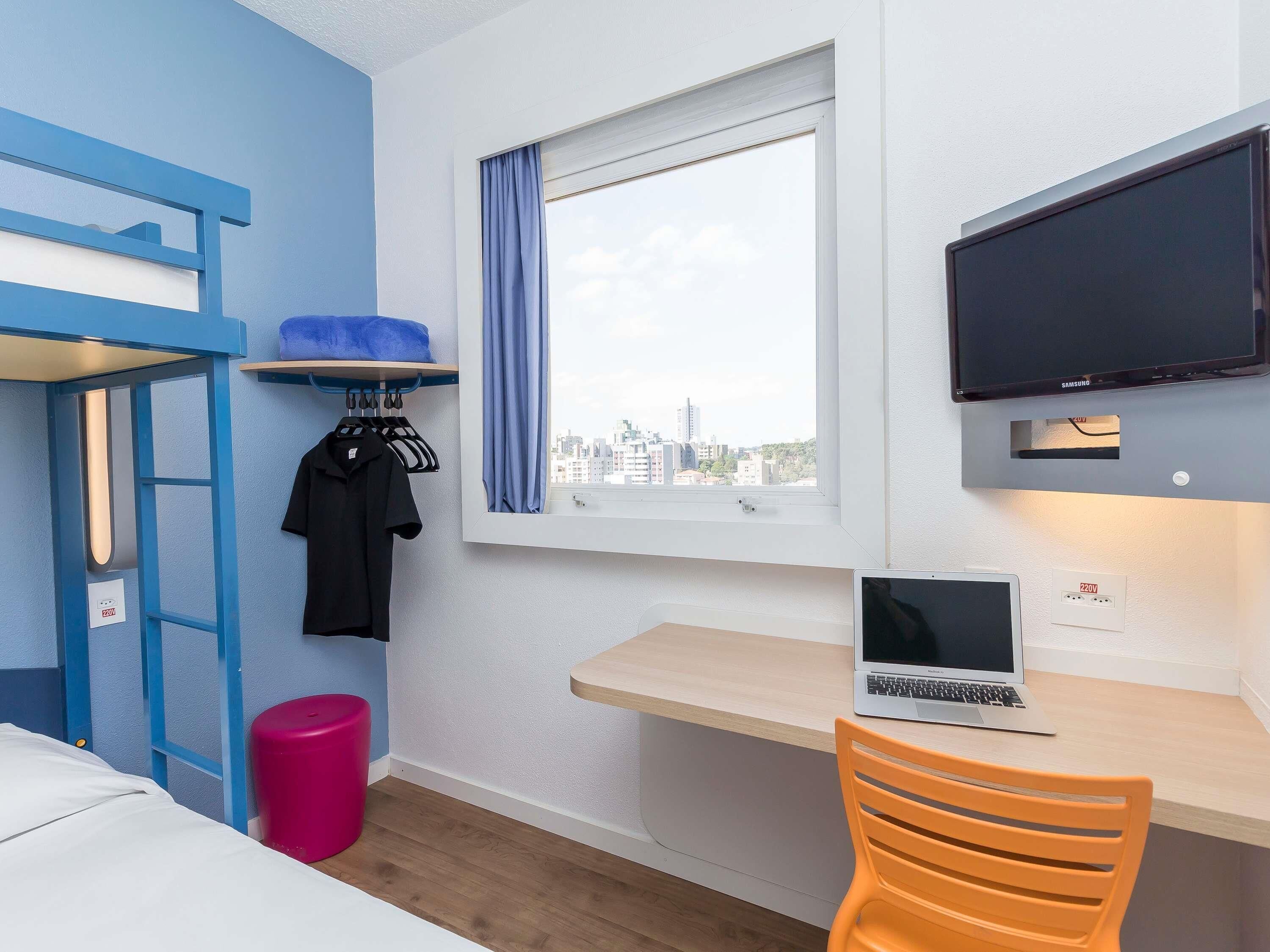 Ett rum med en våningssäng, ett skrivbord med en bärbar dator, en tv och ett fönster med utsikt över staden.