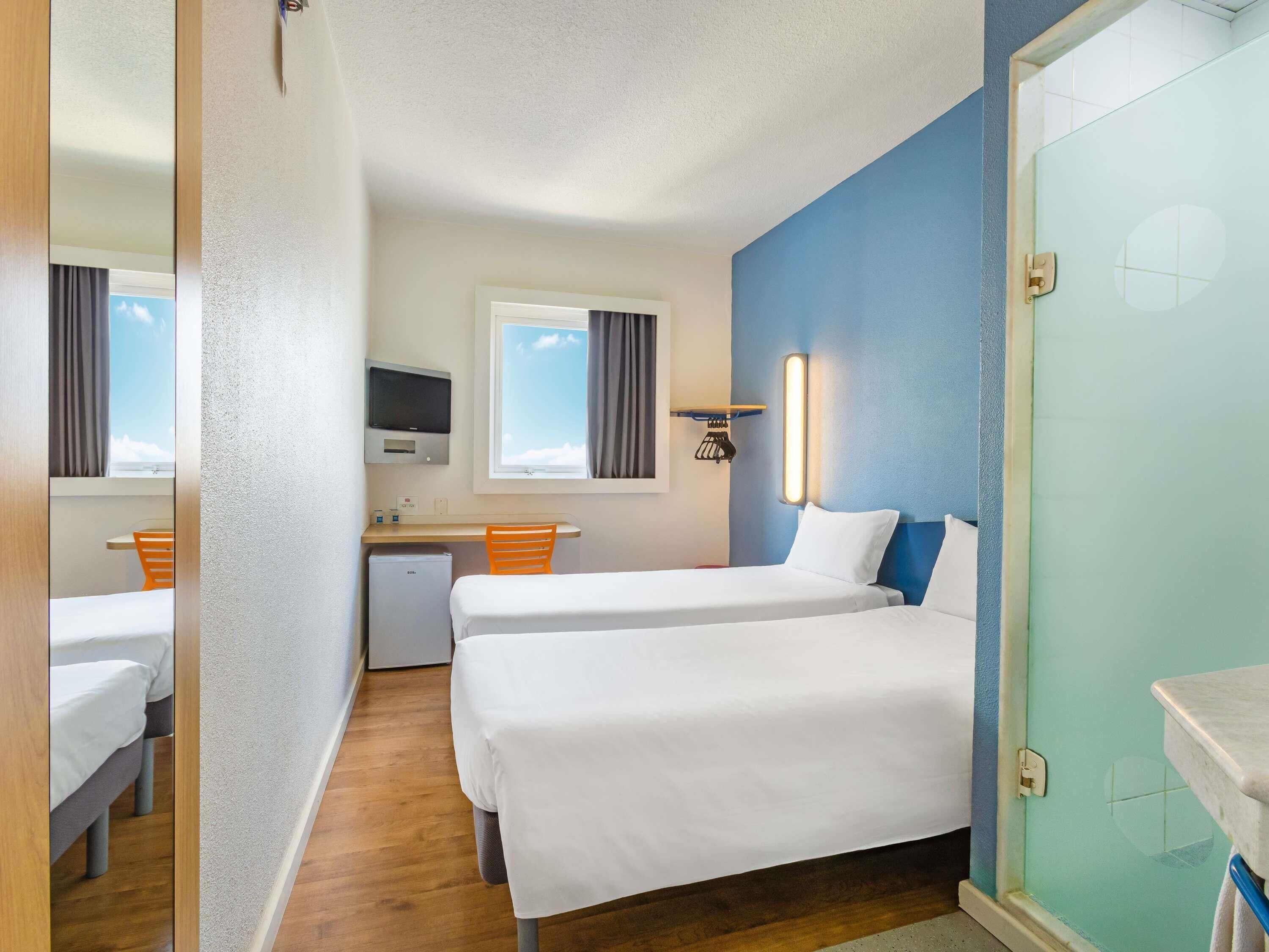 Una habitación de hotel compacta con dos camas, un escritorio, un refrigerador pequeño y una ventana con cortinas.