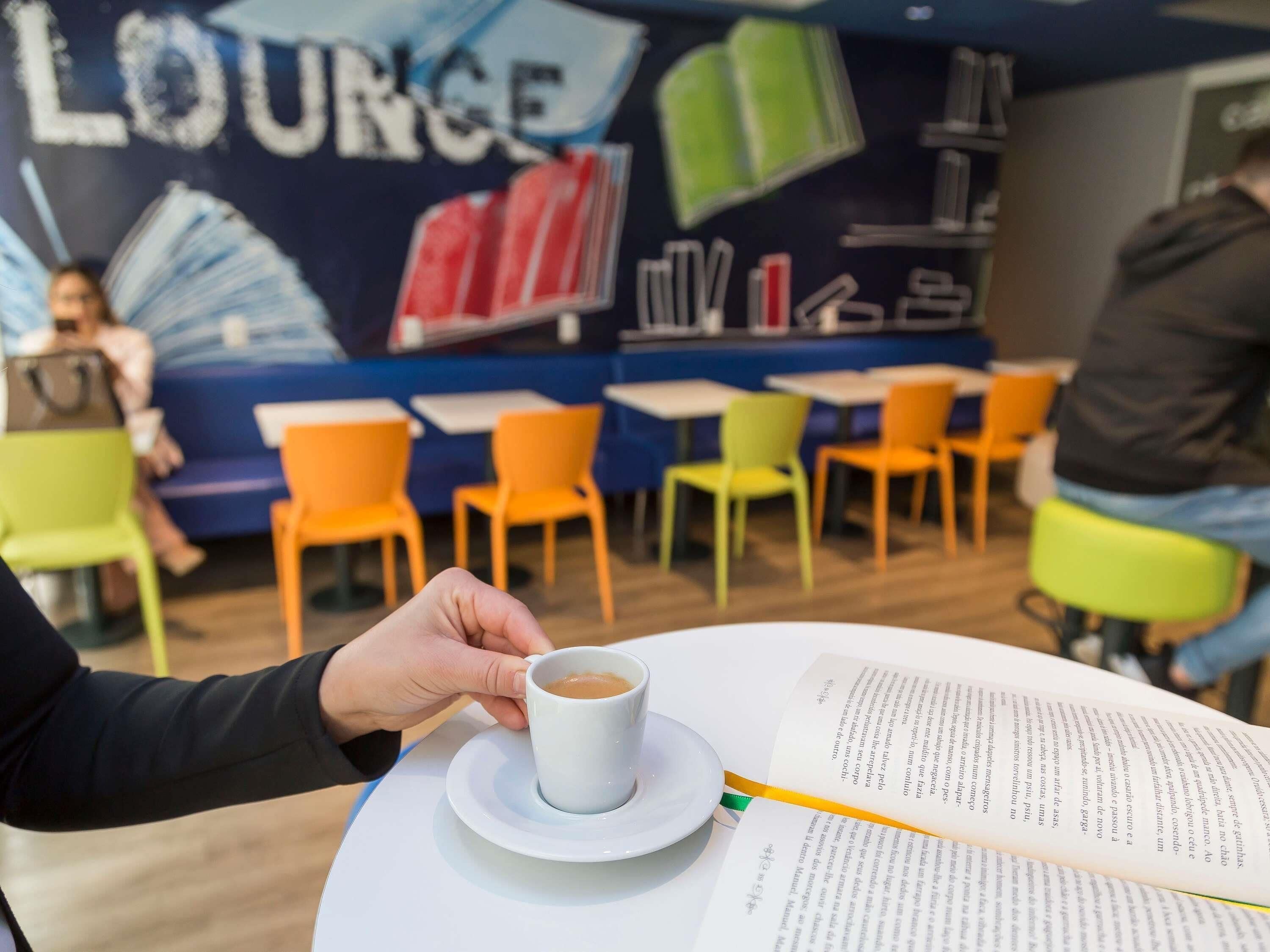 A person holding a cup of coffee, a book, and a menu on a table in a cafe with colorful chairs and a mural in the background.