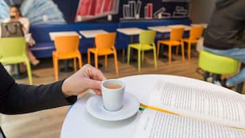 A person holding a cup of coffee, a book, and a menu on a table in a cafe with colorful chairs and a mural in the background.