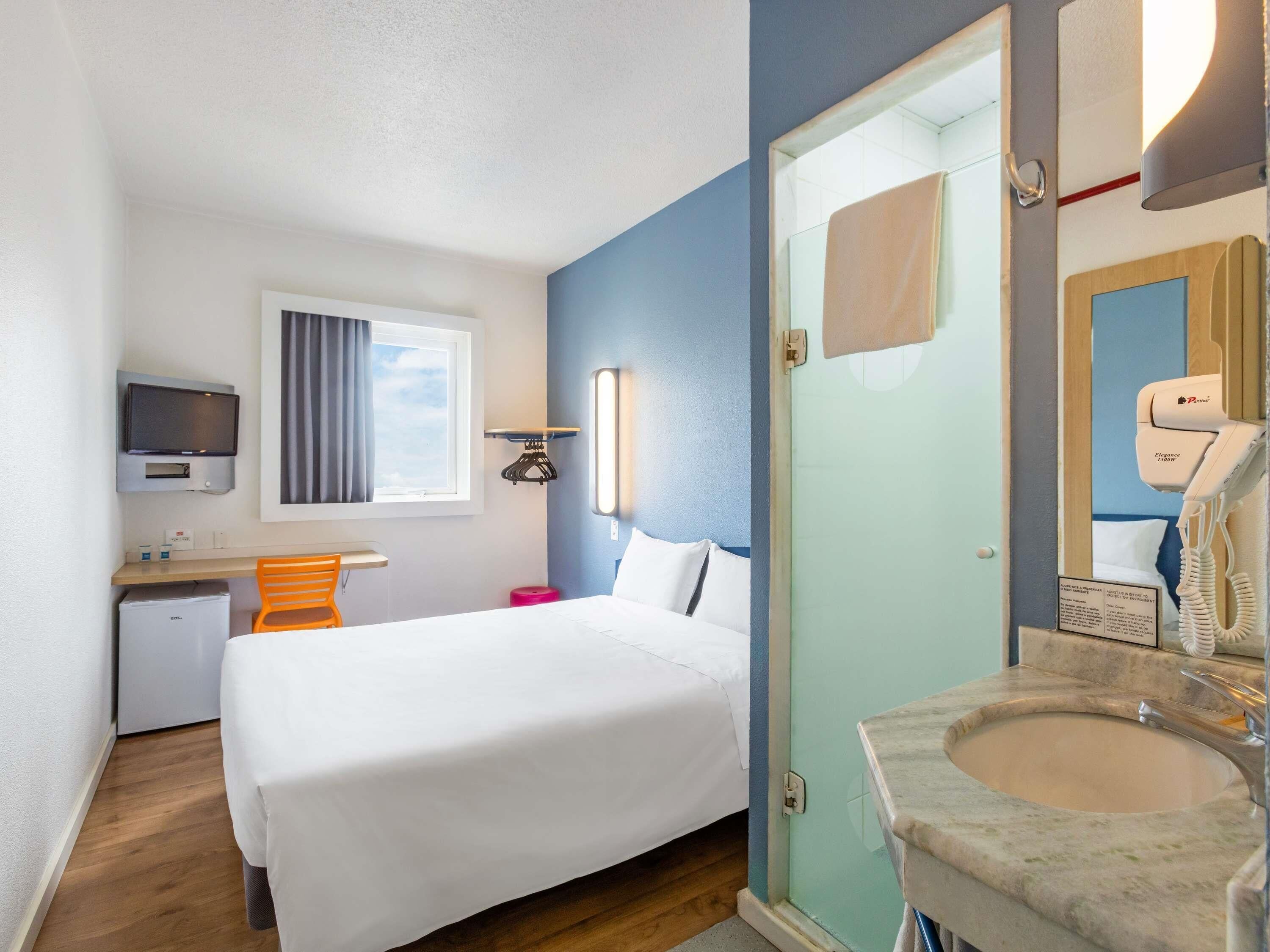 A hotel room with a bed, a desk, a TV, a mirror, and a bathroom with a sink and shower.