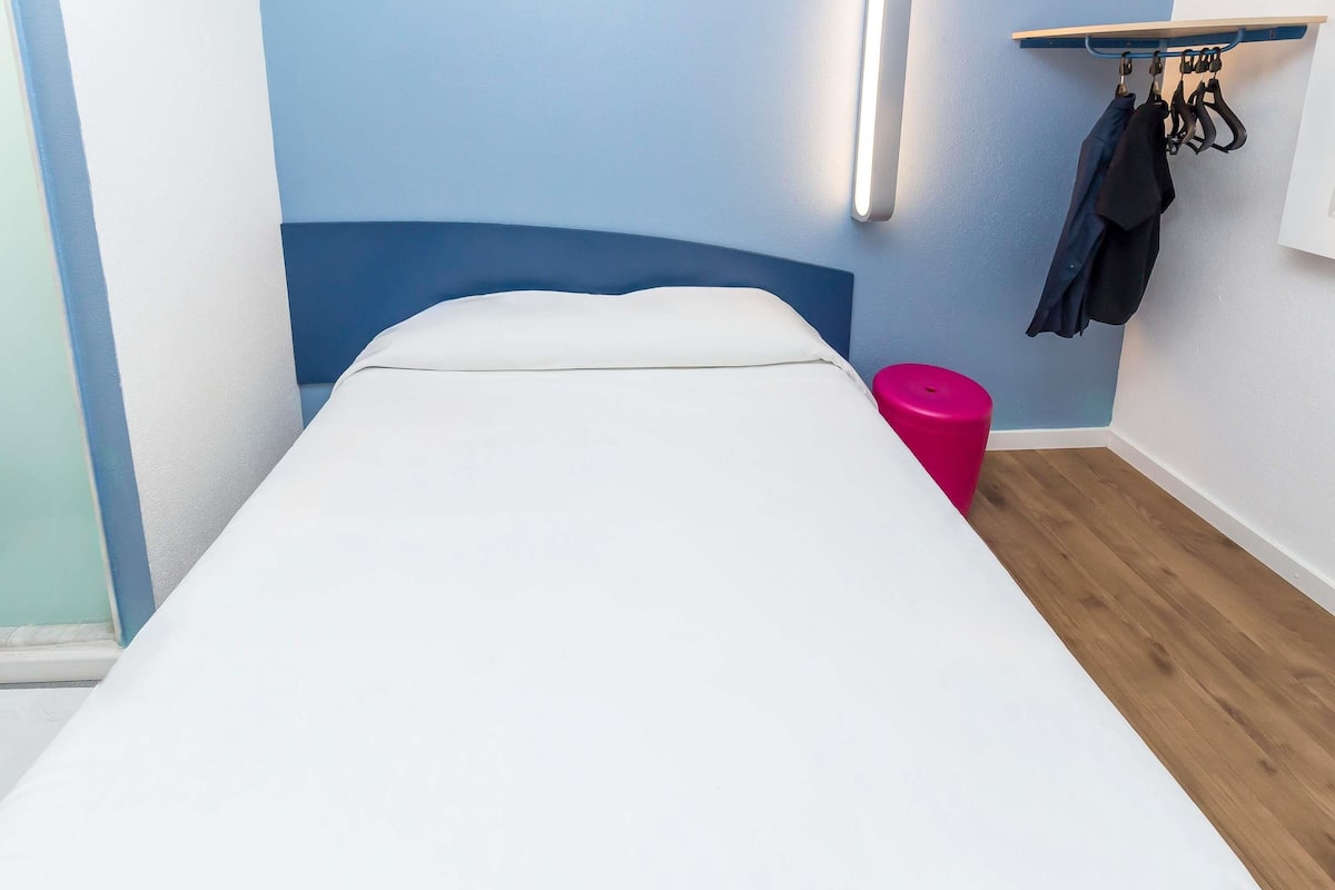 Una cama individual con colchón blanco, cabecera azul y un taburete rosa, además de una lámpara fijada a la pared.