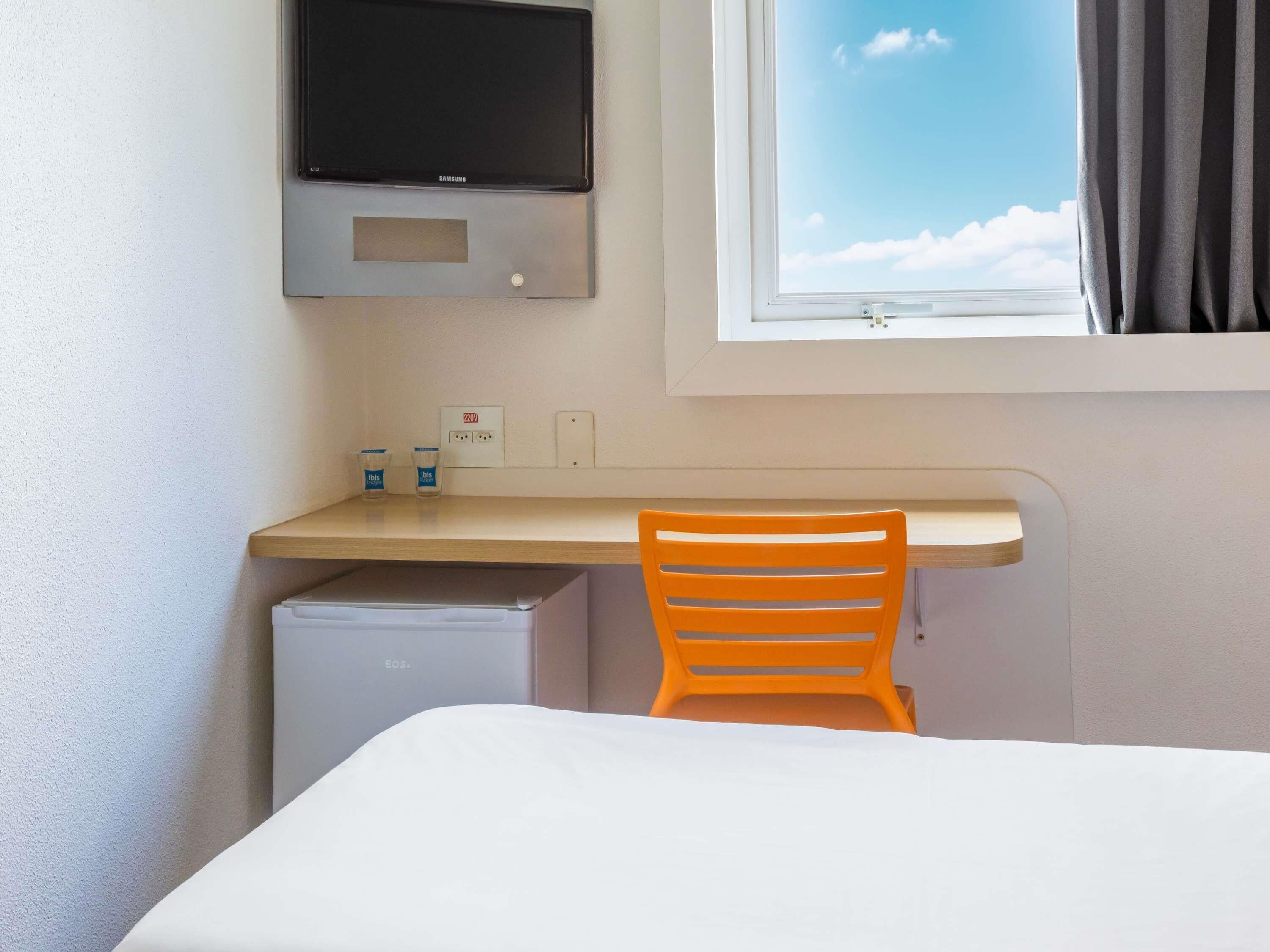 Une chambre d’hôtel compacte comprenant un lit, un bureau avec un écran d’ordinateur, un petit réfrigérateur et une fenêtre donnant sur le ciel.
