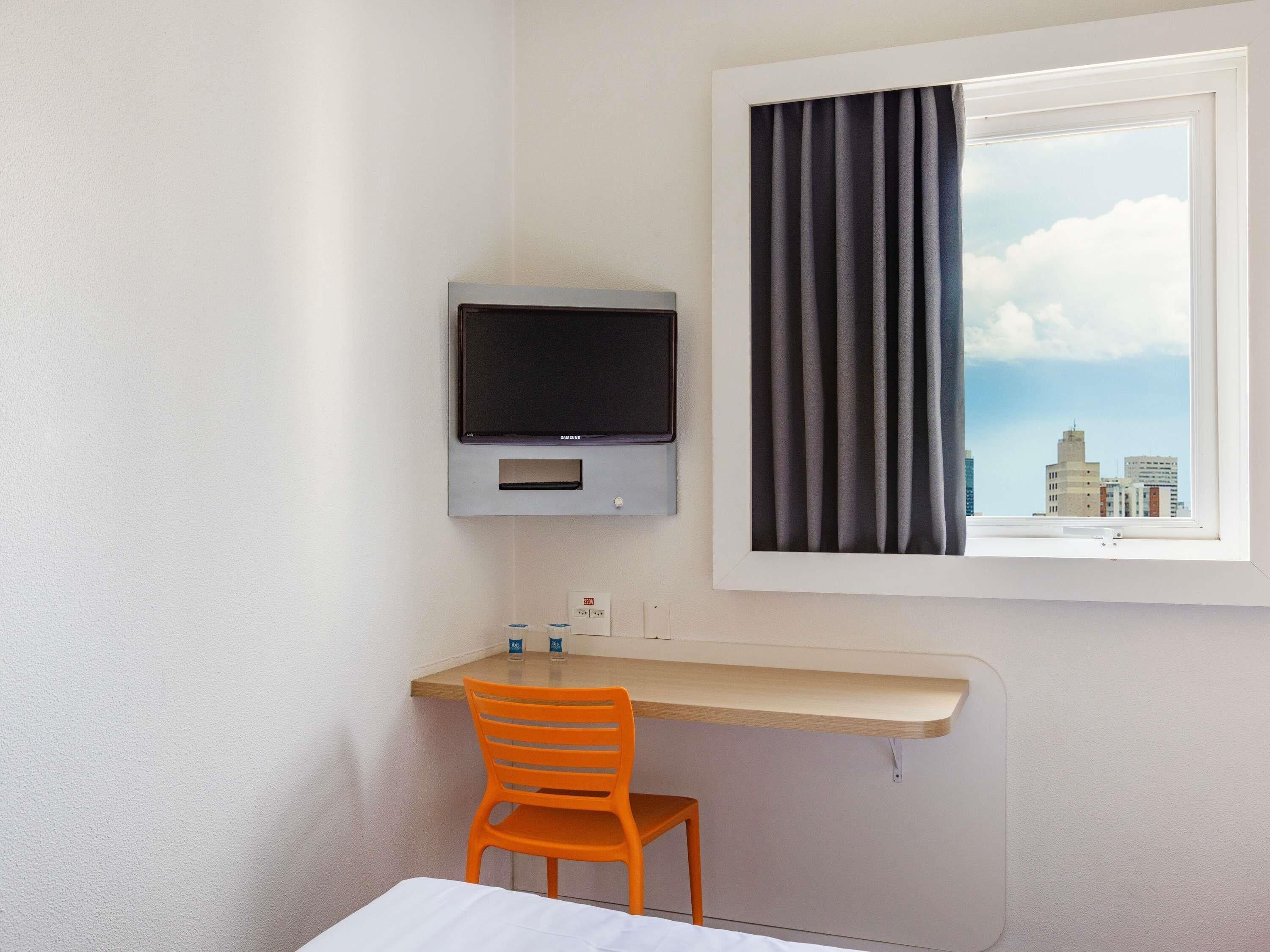 Habitación de hotel con una cama, un escritorio con silla, un televisor montado en la pared y una ventana con cortinas.