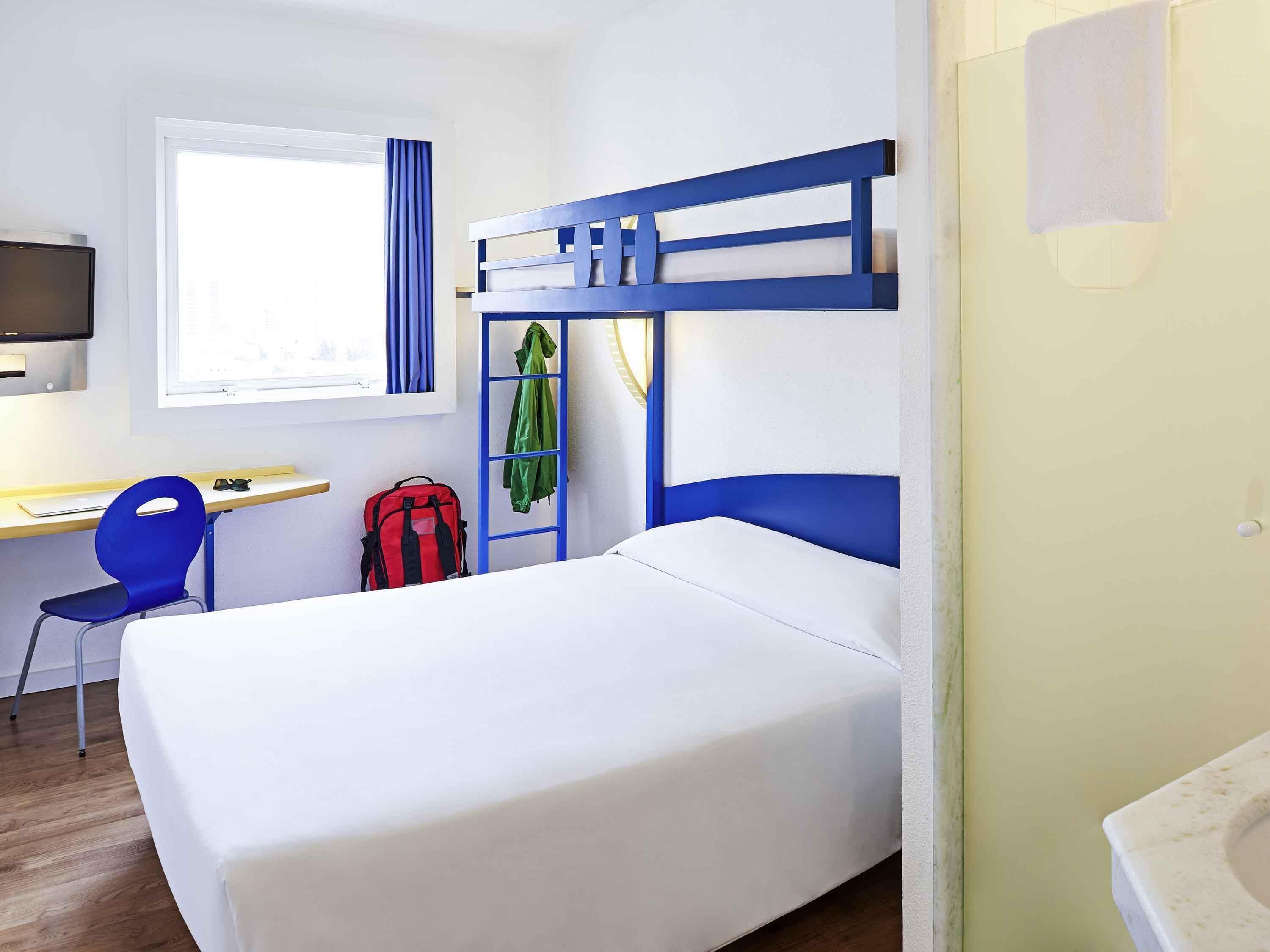 A room with a bunk bed, a desk with a chair, a TV mounted on the wall, and a window with curtains.