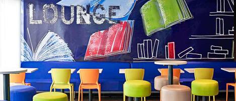 Zona relax con poltrone e tavolini colorati, un divano blu e una parete decorata con un'illustrazione di una libreria.