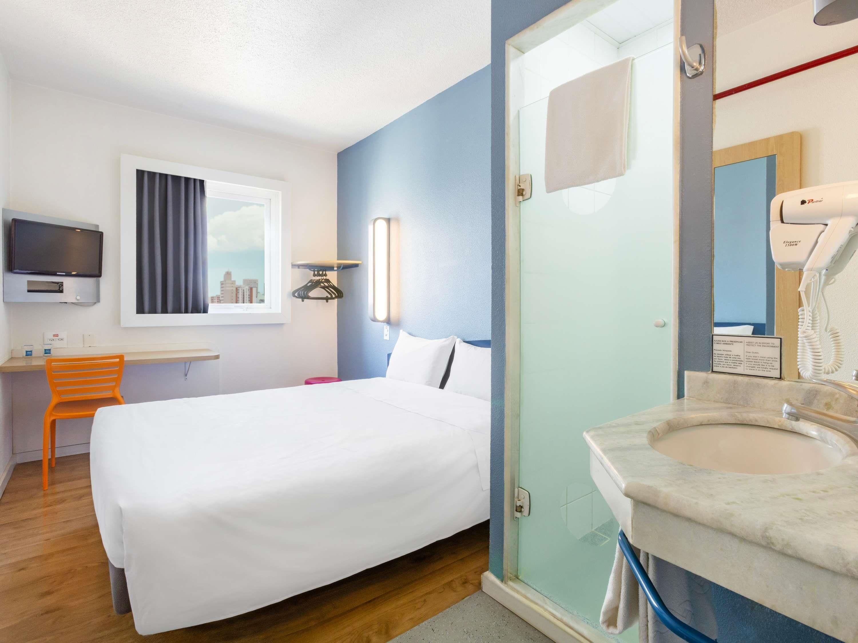 A hotel room with a bed, a desk, a TV, a window with curtains, a shower area, and a bathroom with a sink and mirror.