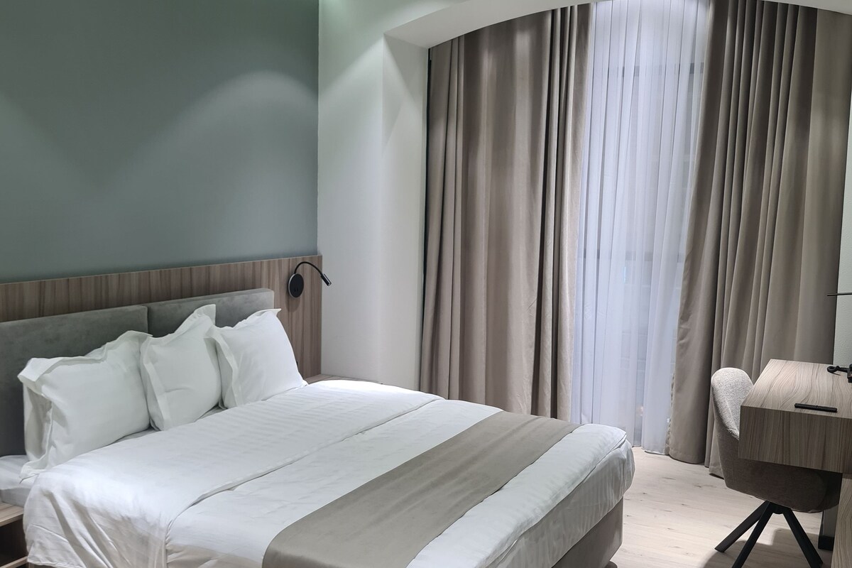 A modern bedroom with a large bed, a desk, and a curved window with curtains.