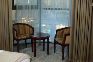 Une chambre d’hôtel avec un lit, deux fauteuils, une petite table et une vue à travers des rideaux légers.