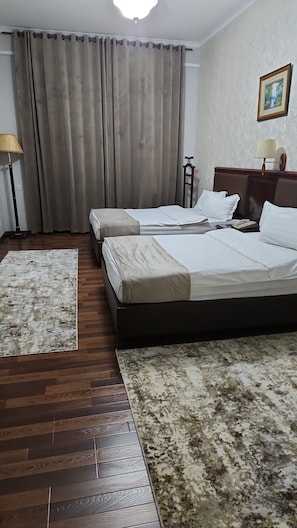 Une chambre d’hôtel avec deux lits, un plancher en bois, une grande fenêtre avec des rideaux, une lampe sur pied et un tableau encadré au mur.