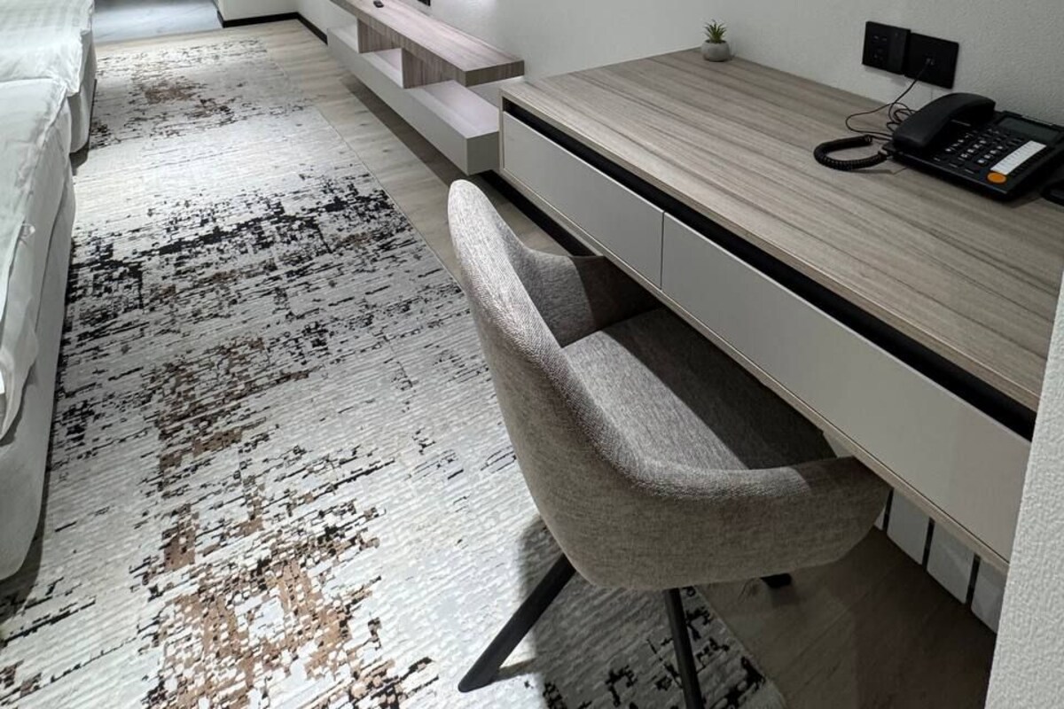 A modern hotel room with a wooden desk, a flat-screen TV mounted on the wall, a telephone, and a patterned rug.