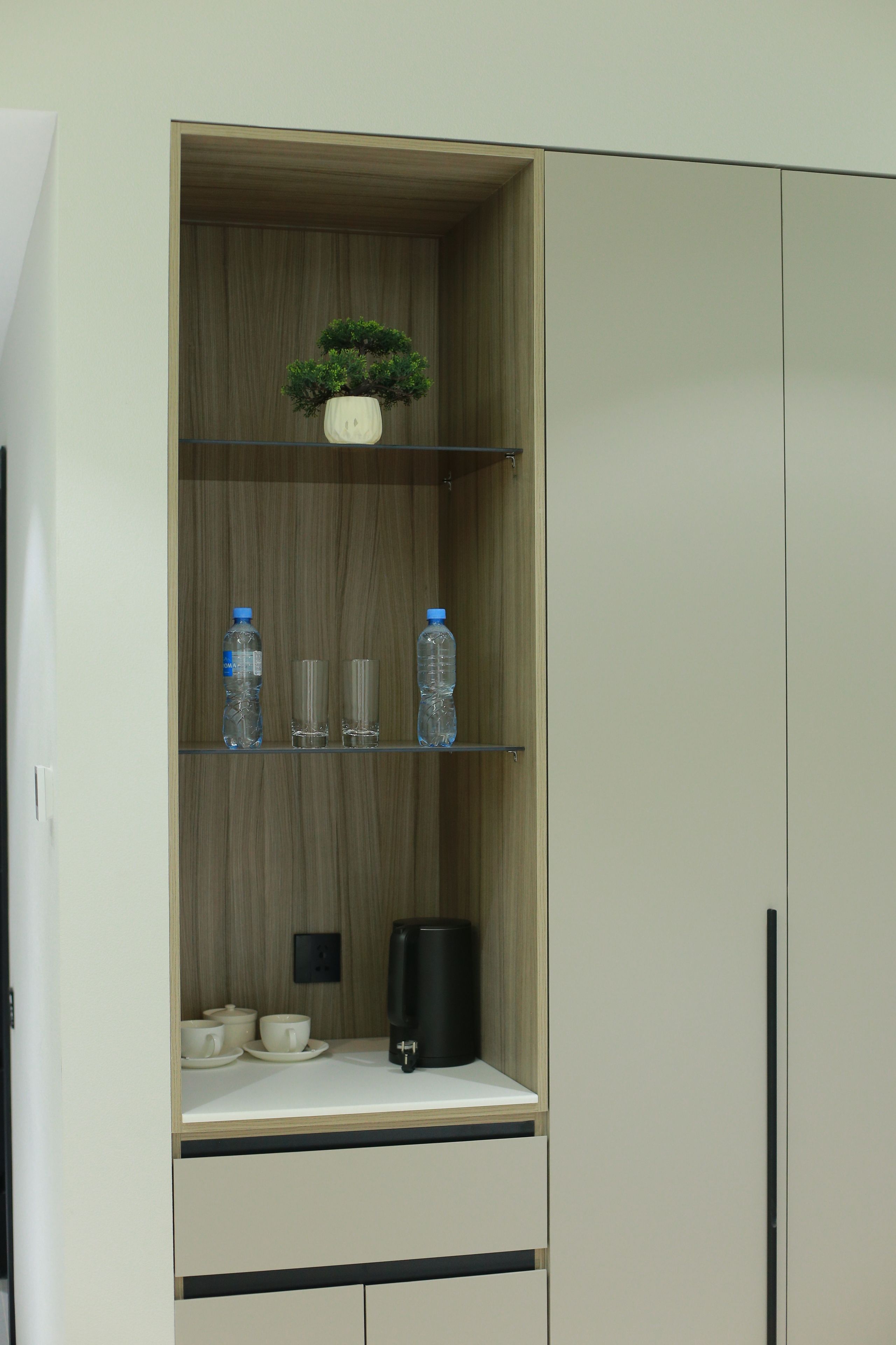 A cabinet with a plant, water bottles, glasses, a coffee maker, and cups.