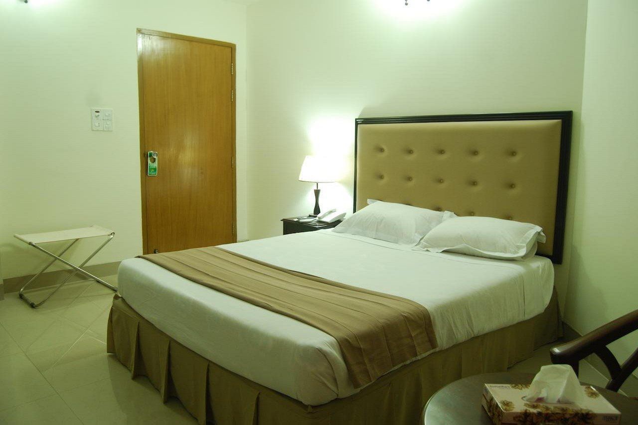 Quarto de hotel com cama, mesa de cabeceira, abajur, cadeira dobrável e porta.