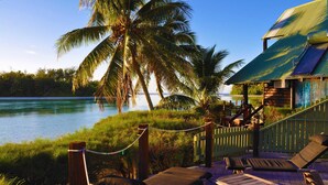 A wooden deck with lounge chairs overlooking a body of water, palm trees, and a building with a green roof. - Sokala Villas (Rarotonga)