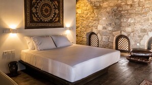 Un lit avec une tête de lit, deux lampes fixées au mur, un mur en pierre et une tapisserie décorative.