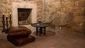 A stone-walled room with a fireplace, two wicker chairs, a round table, and a brown leather ottoman.