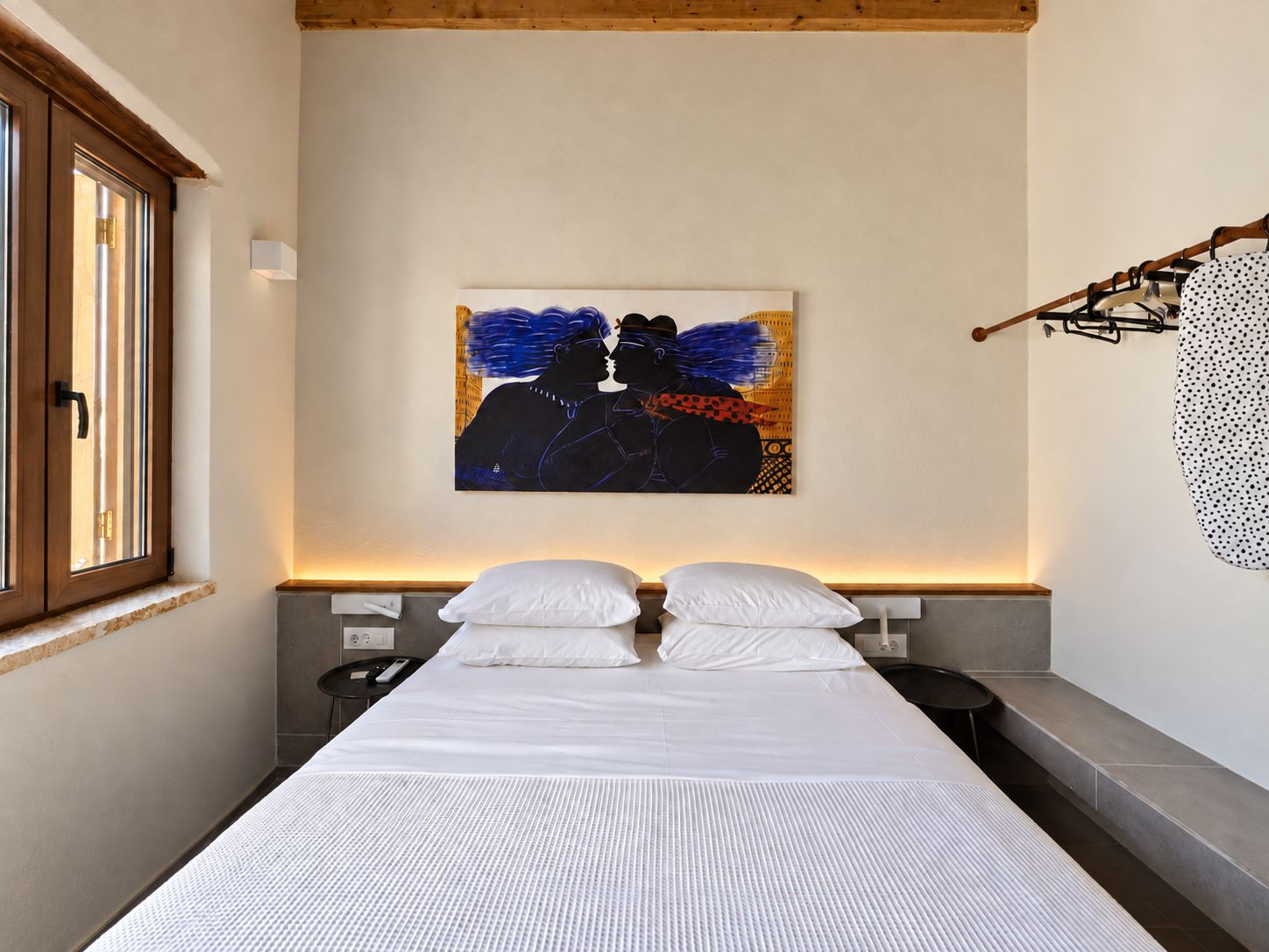 A bedroom with a bed, a painting, a window, and a coat rack.
