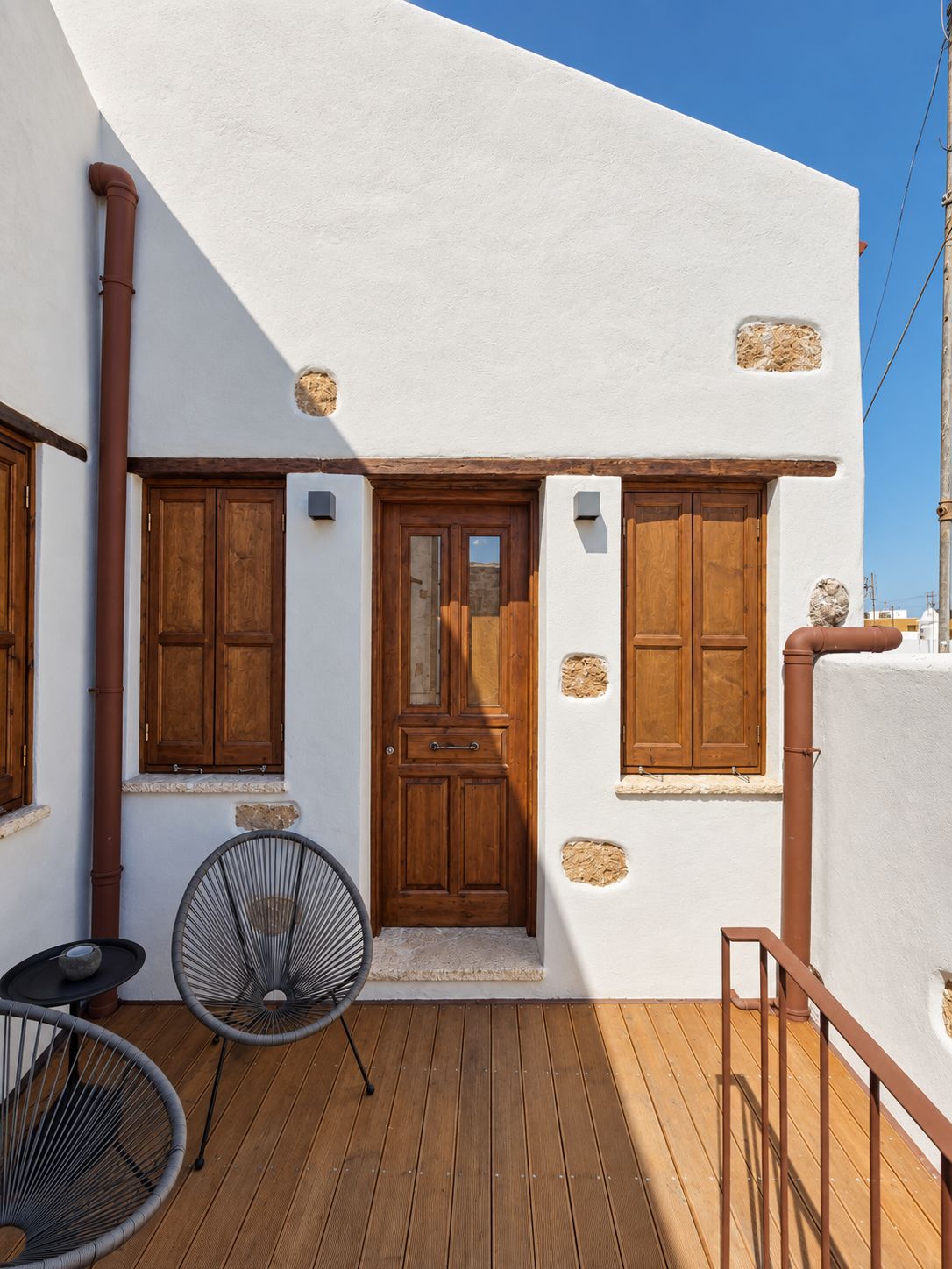 A white building with a wooden door, two windows with shutters, a wooden deck, and a metal chair.