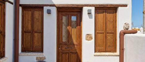 A white building with a wooden door, two windows with shutters, a wooden deck, and a metal chair.