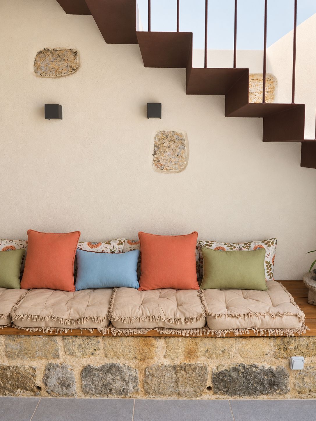 Un intérieur moderne avec un mur en pierre, un banc rembourré agrémenté de coussins colorés et un escalier doté d’une rampe en métal.