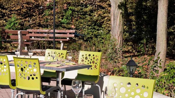 Une terrasse extérieure avec des tables et des chaises, entourée d’arbres et d’une haie.