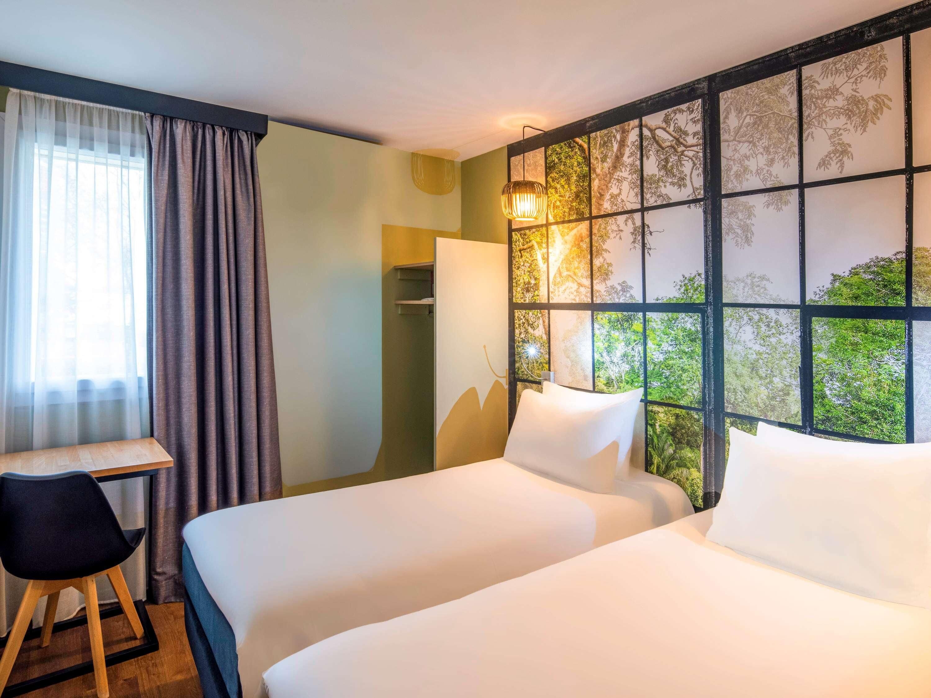 A hotel room with two beds, a desk, a chair, and a view through a window with a tree pattern.