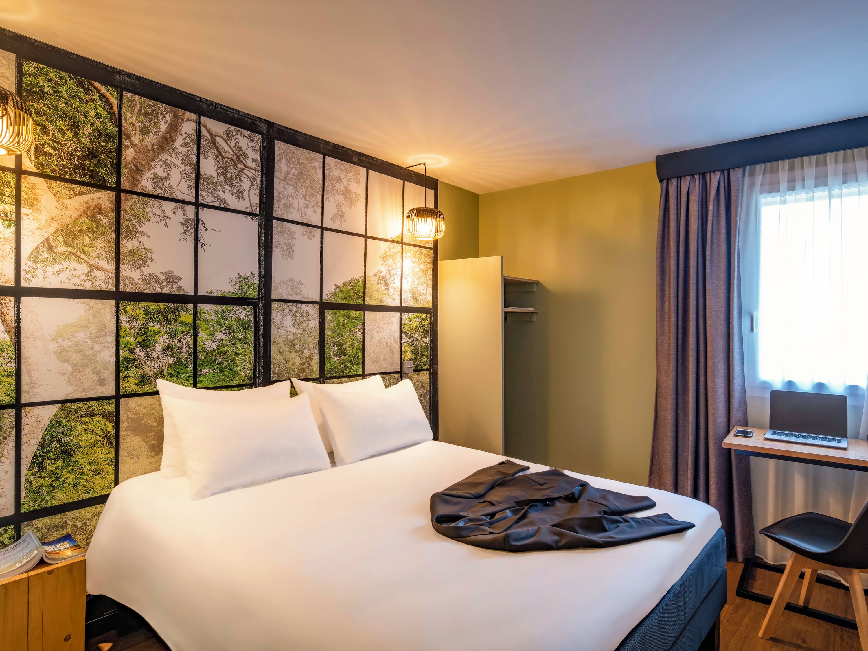 A hotel room with a bed, a desk with a laptop, and a view of trees through a window.