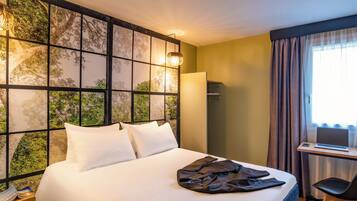 A hotel room with a bed, a desk with a laptop, and a view of trees through a window.