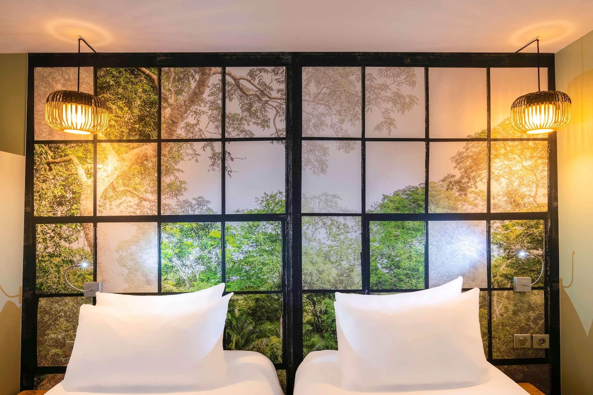 Une chambre avec deux lits, une grande fenêtre donnant sur la forêt et deux lampes fixées au mur.