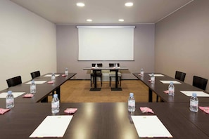 Sala de conferência com tela grande, mesas compridas, cadeiras, garrafas de água e papéis.
