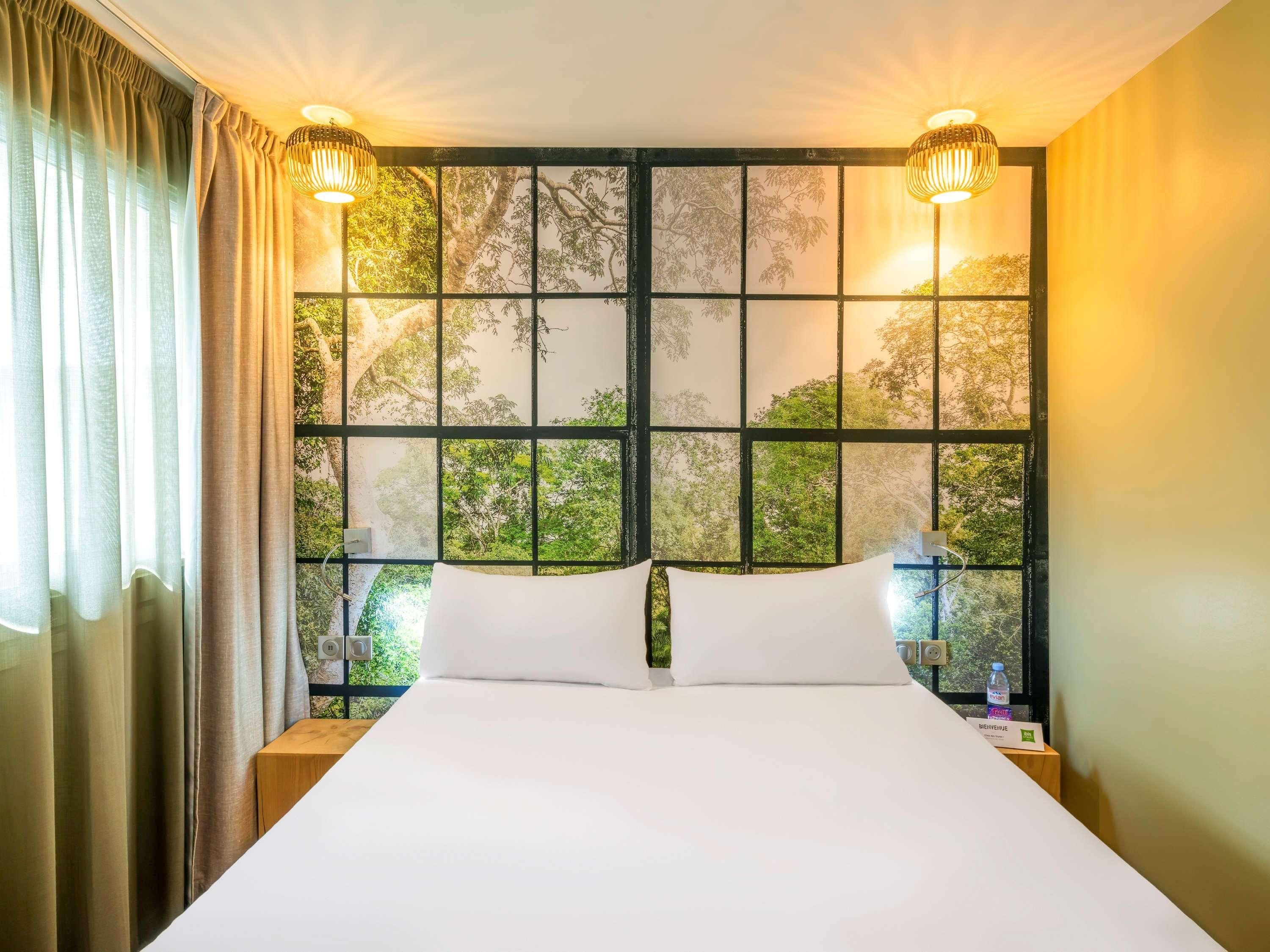 Habitación de hotel con una cama grande, dos lámparas de noche, un ventanal con vista a árboles y una mesita de noche con una botella.