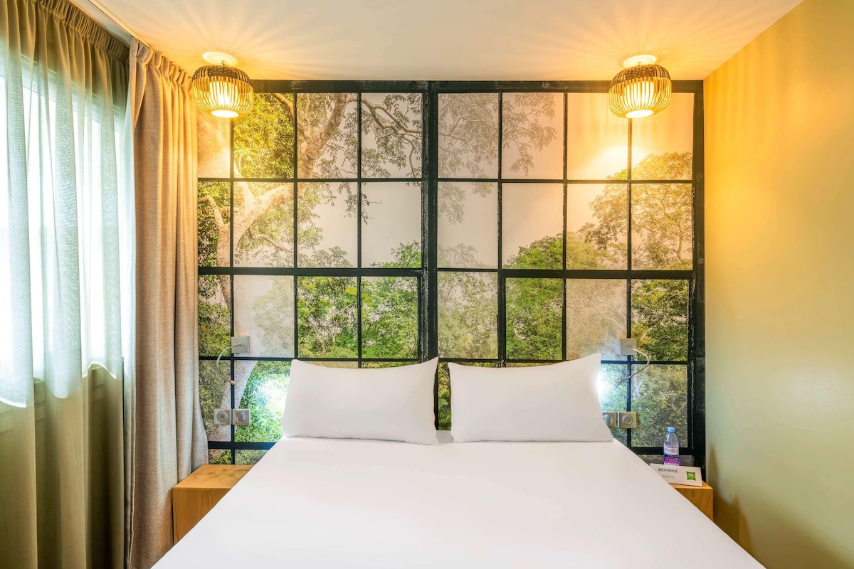 Une chambre d’hôtel avec un grand lit, deux lampes de chevet, une fenêtre donnant sur des arbres et une table de chevet avec une bouteille.