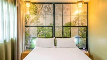Habitación de hotel con una cama grande, dos lámparas de noche, un ventanal con vista a árboles y una mesita de noche con una botella.