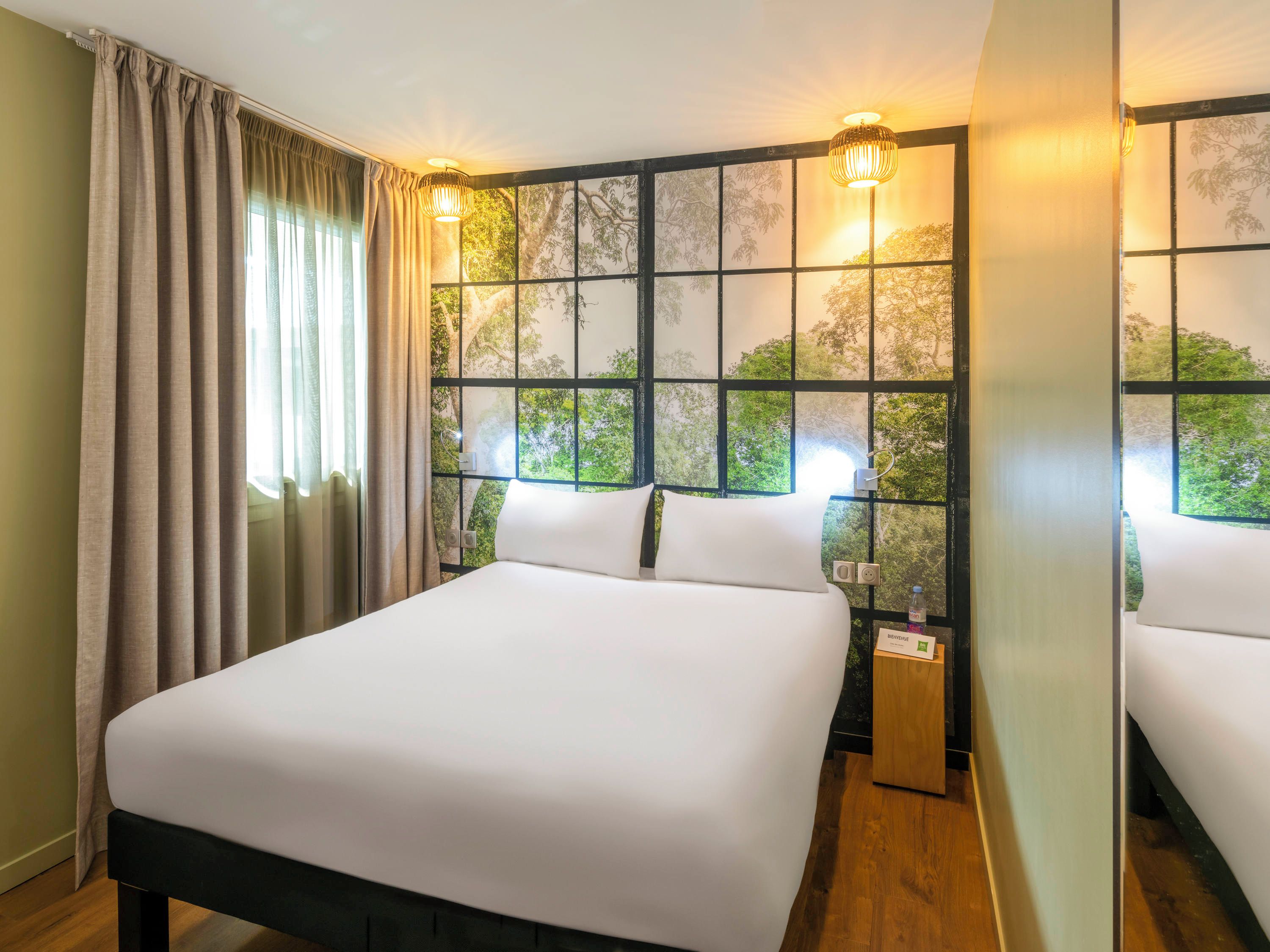 A hotel room with a large bed, two bedside tables, a window with a view of trees, and two wall-mounted lamps.