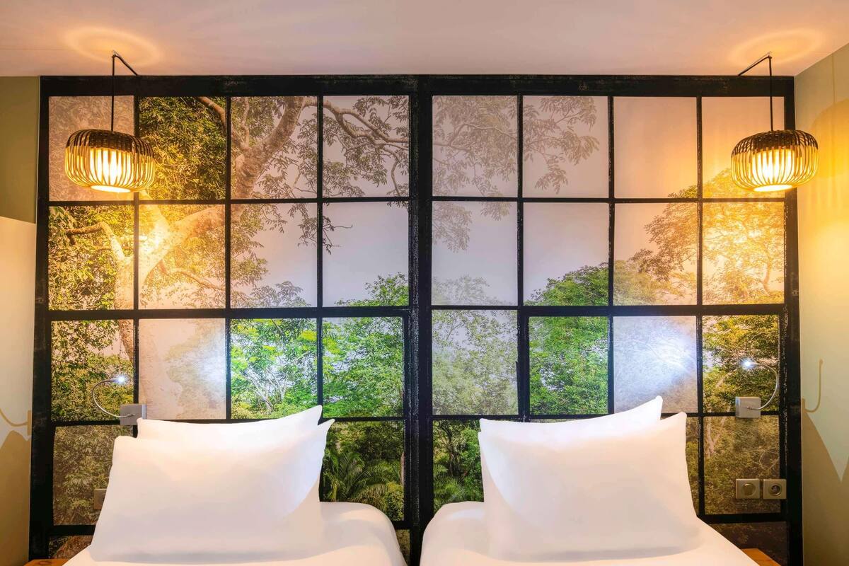 Een kamer met twee bedden, een groot raam met uitzicht op bomen, en twee wandlampen.