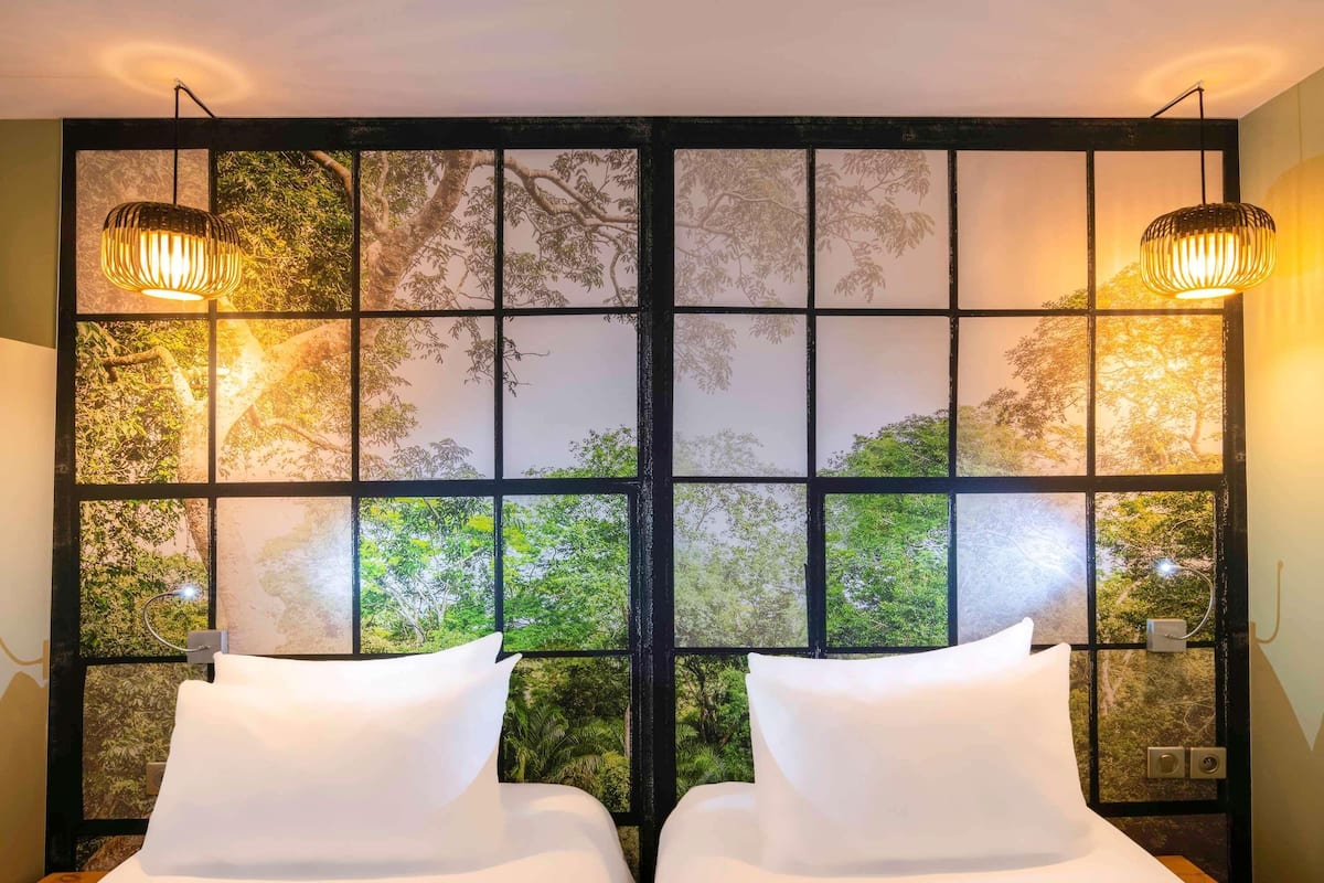 A room with two beds, a large window with a view of trees, and two wall-mounted lamps.