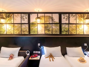 Une chambre d’hôtel avec deux lits, une grande fenêtre donnant sur la verdure et un luminaire fixé au mur.