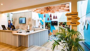Un espace d’accueil moderne avec un comptoir de réception en bois, un écran de télévision et des panneaux indicateurs pour un restaurant et pour la ville de Paris.