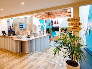 Un espace d’accueil moderne avec un comptoir de réception en bois, un écran de télévision et des panneaux indicateurs pour un restaurant et pour la ville de Paris.