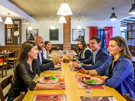 Un grupo de personas cenando en un restaurante con una larga mesa de madera, luces colgantes blancas y un muro de ladrillo al fondo.