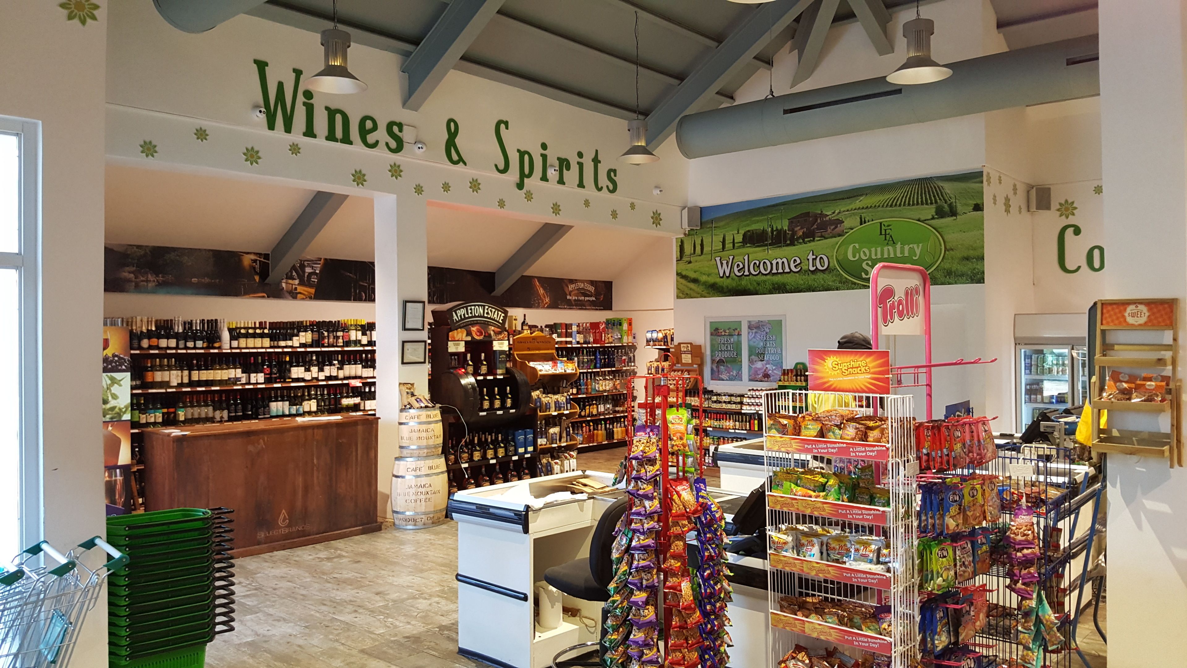 A store interior with shelves stocked with various products, a section labeled "Wines & Spirits," and a "Welcome to Country" sign.