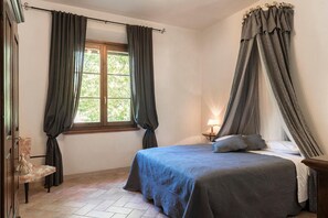 Une chambre à coucher avec un lit, une fenêtre avec des rideaux, une chaise et une table de chevet avec une lampe.