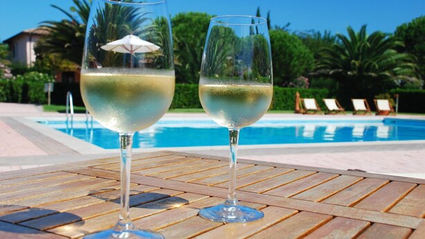 Two glasses of white wine on a wooden table by a poolside with lounge chairs and greenery in the background. - Agriturismo Villa Toscana (Campiglia Marittima)