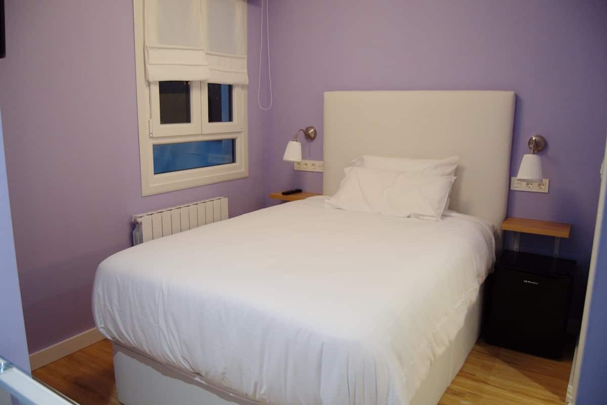 Un lit bien fait dans une pièce aux murs violets, une fenêtre avec des stores blancs, une table de chevet avec une lampe et une valise posée sur le sol.