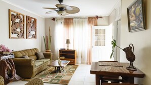 A living room with a sofa, coffee table, and dining table. There is a ceiling fan, a window with curtains, and a door.