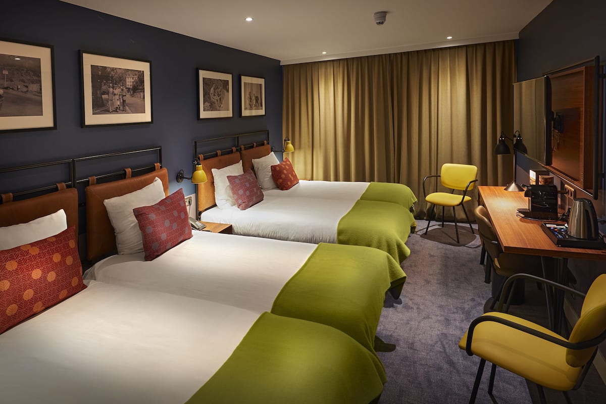 A hotel room with two beds, a desk, chairs, a TV, and framed pictures on the wall.