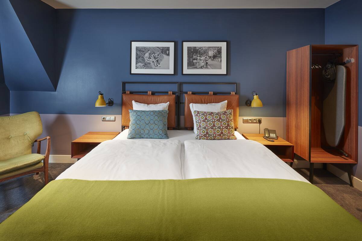 A double bed with two pillows, a wooden nightstand with a lamp, a wooden wardrobe, and two framed pictures on the wall.